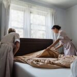 Two housekeepers making a bed in a sunlit bedroom, showcasing cleanliness and attention to detail.